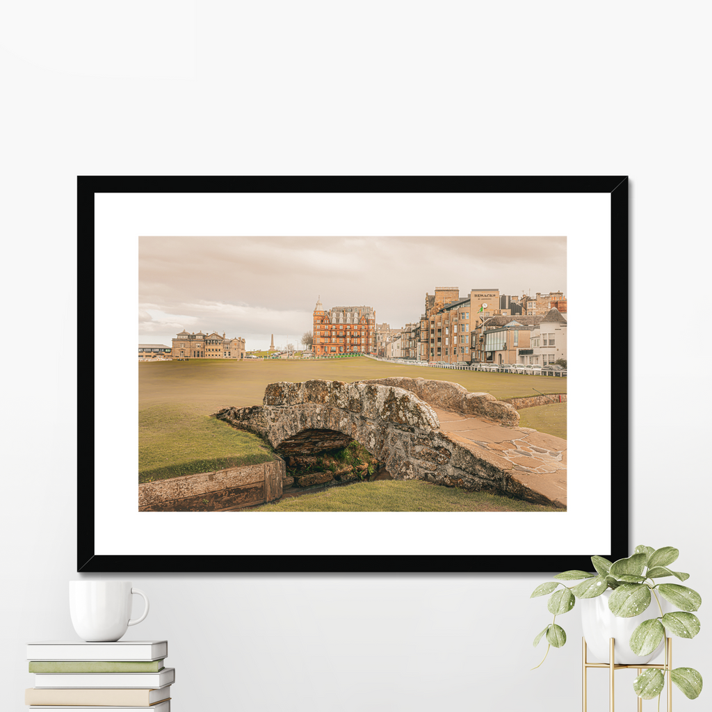 A fine art print of the St Andrews Swilcan Bridge displayed in a modern black frame with a white mount, styled on a shelf next to white books and a small potted plant.