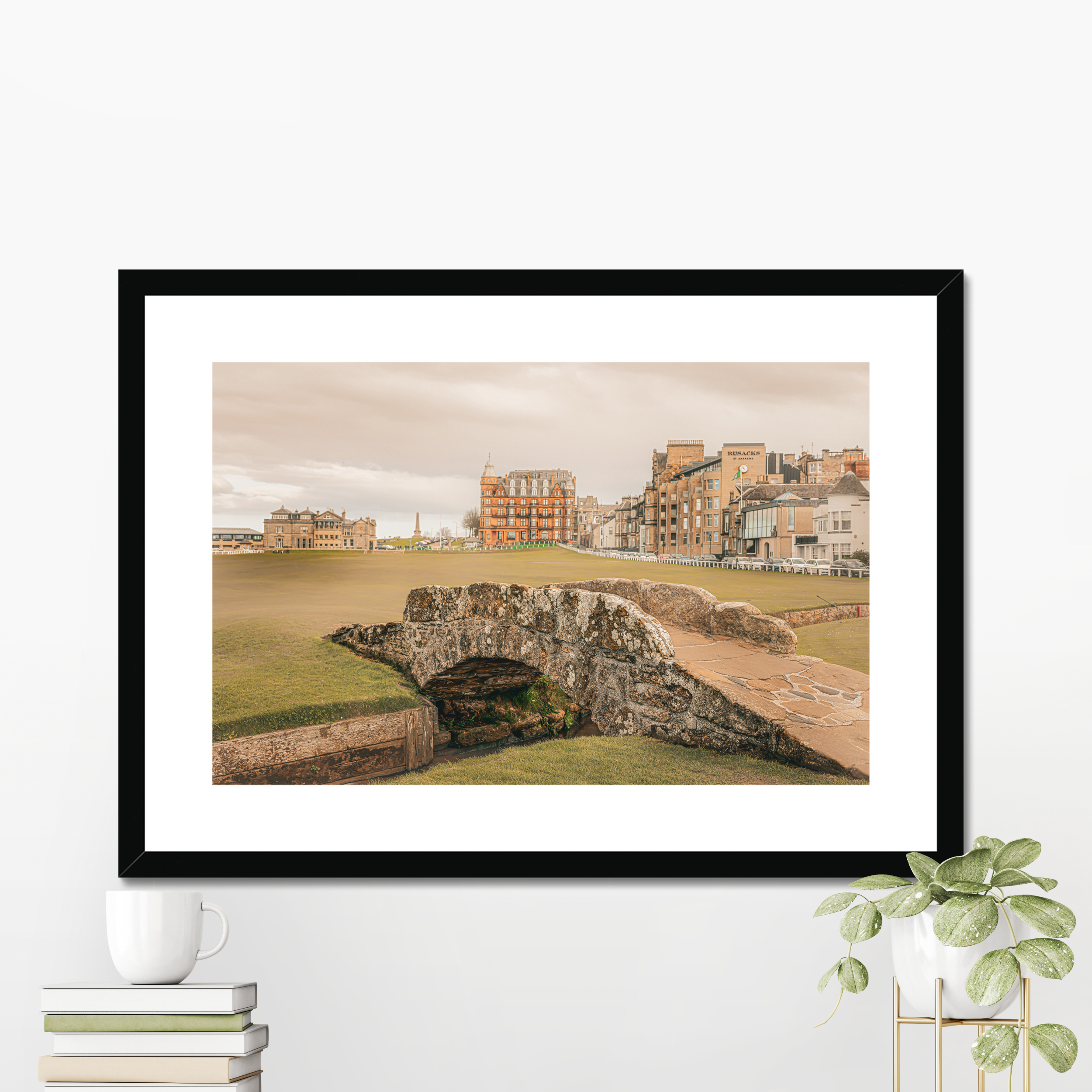 A fine art print of the St Andrews Swilcan Bridge displayed in a modern black frame with a white mount, styled on a shelf next to white books and a small potted plant.