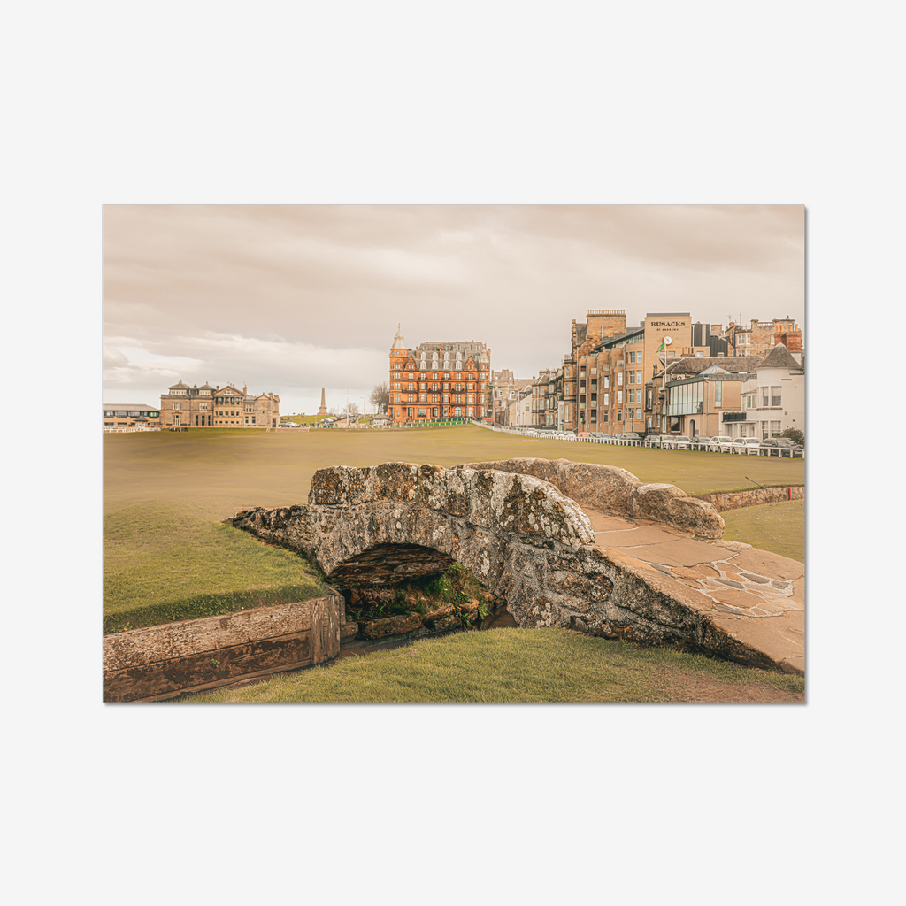 A wide-angle landscape photograph of the historic stone Swilcan Bridge on the 18th fairway of the Old Course at St Andrews, Scotland, under a soft overcast sky with the town's historic buildings in the background.
