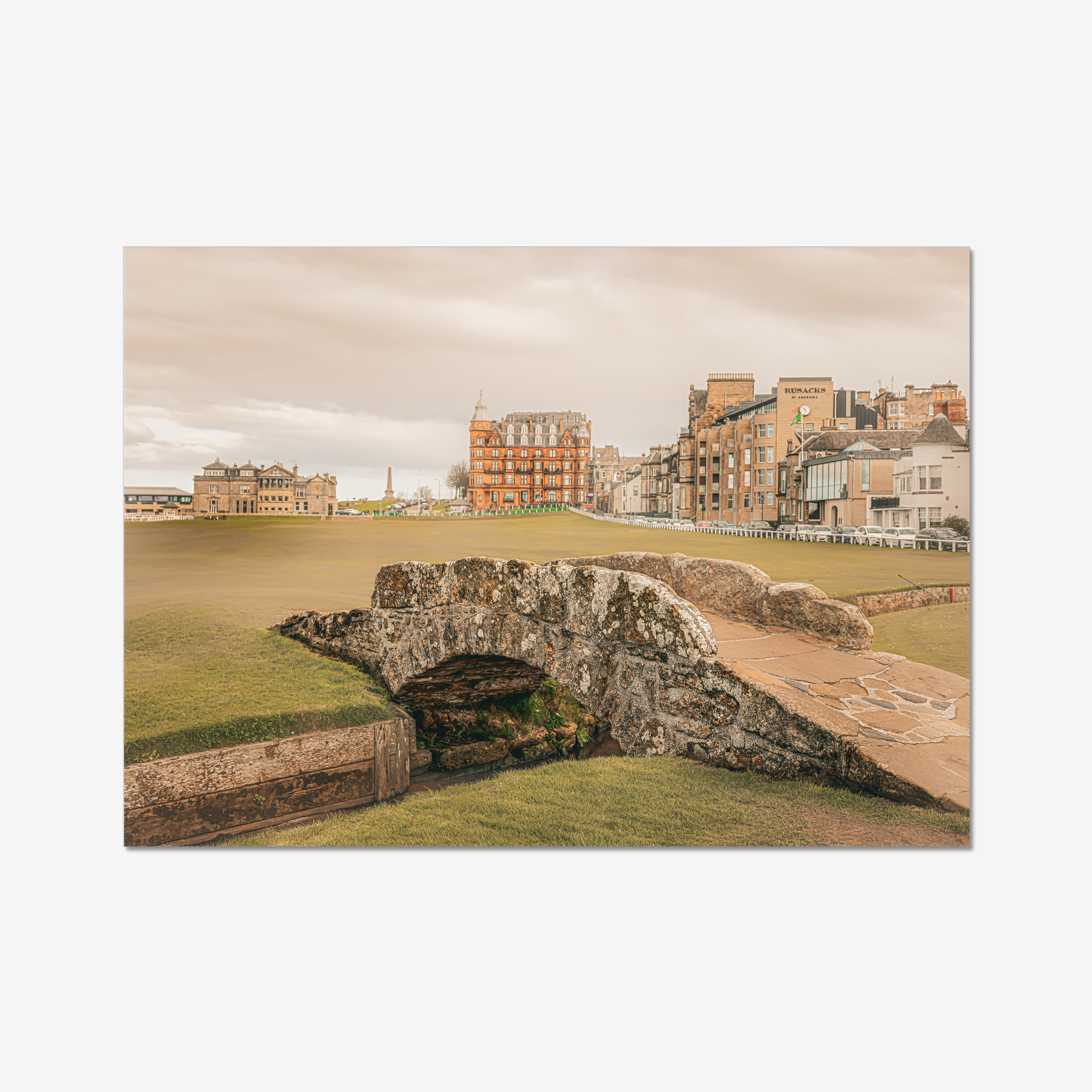 A wide-angle landscape photograph of the historic stone Swilcan Bridge on the 18th fairway of the Old Course at St Andrews, Scotland, under a soft overcast sky with the town's historic buildings in the background.